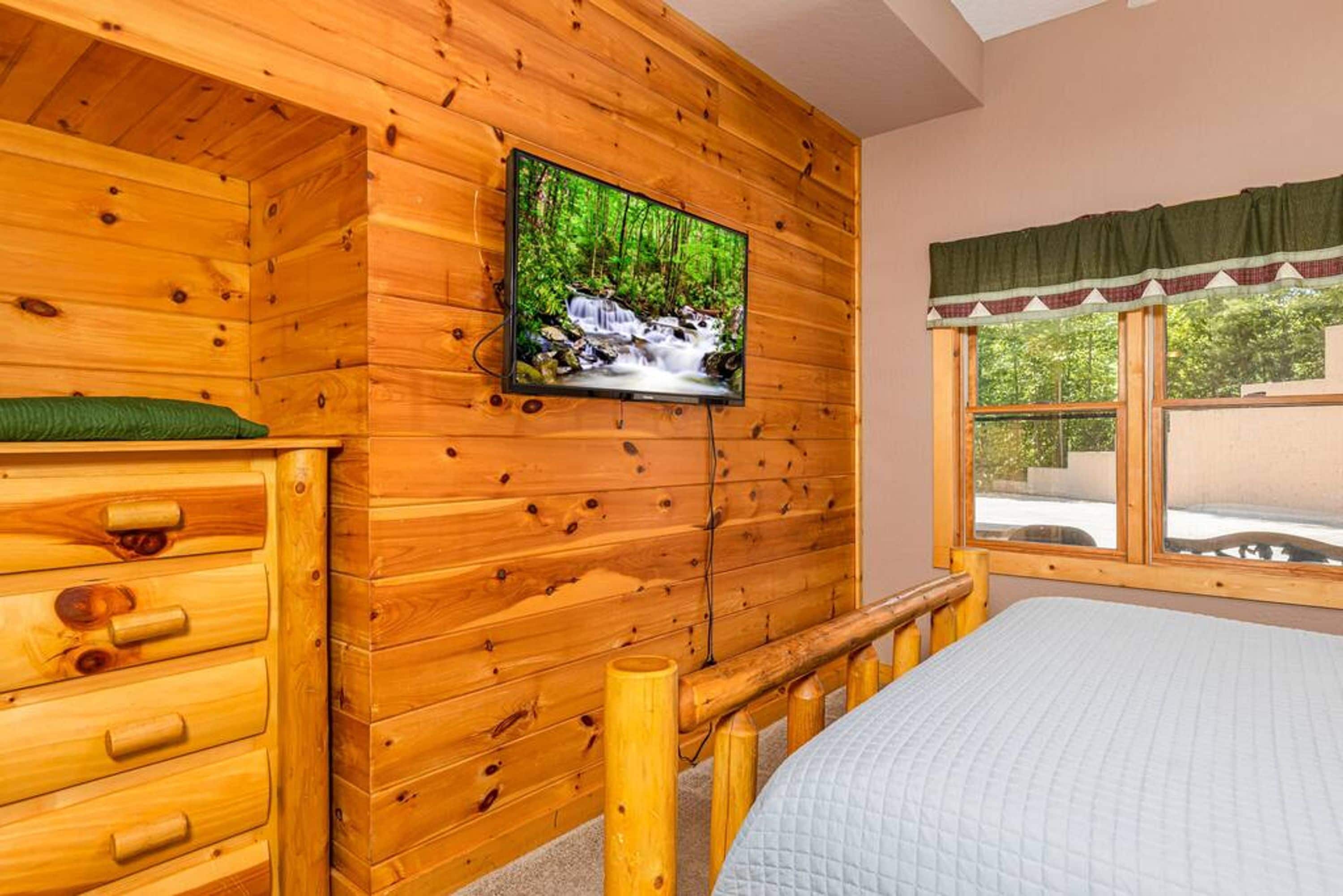 A wooden cabin room with a bed, a TV mounted on the wall, and a window with a view of greenery.