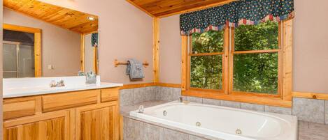 Une salle de bain avec des meubles en bois, une grande baignoire et une fenêtre agrémentée d’une lambrequin à motifs.