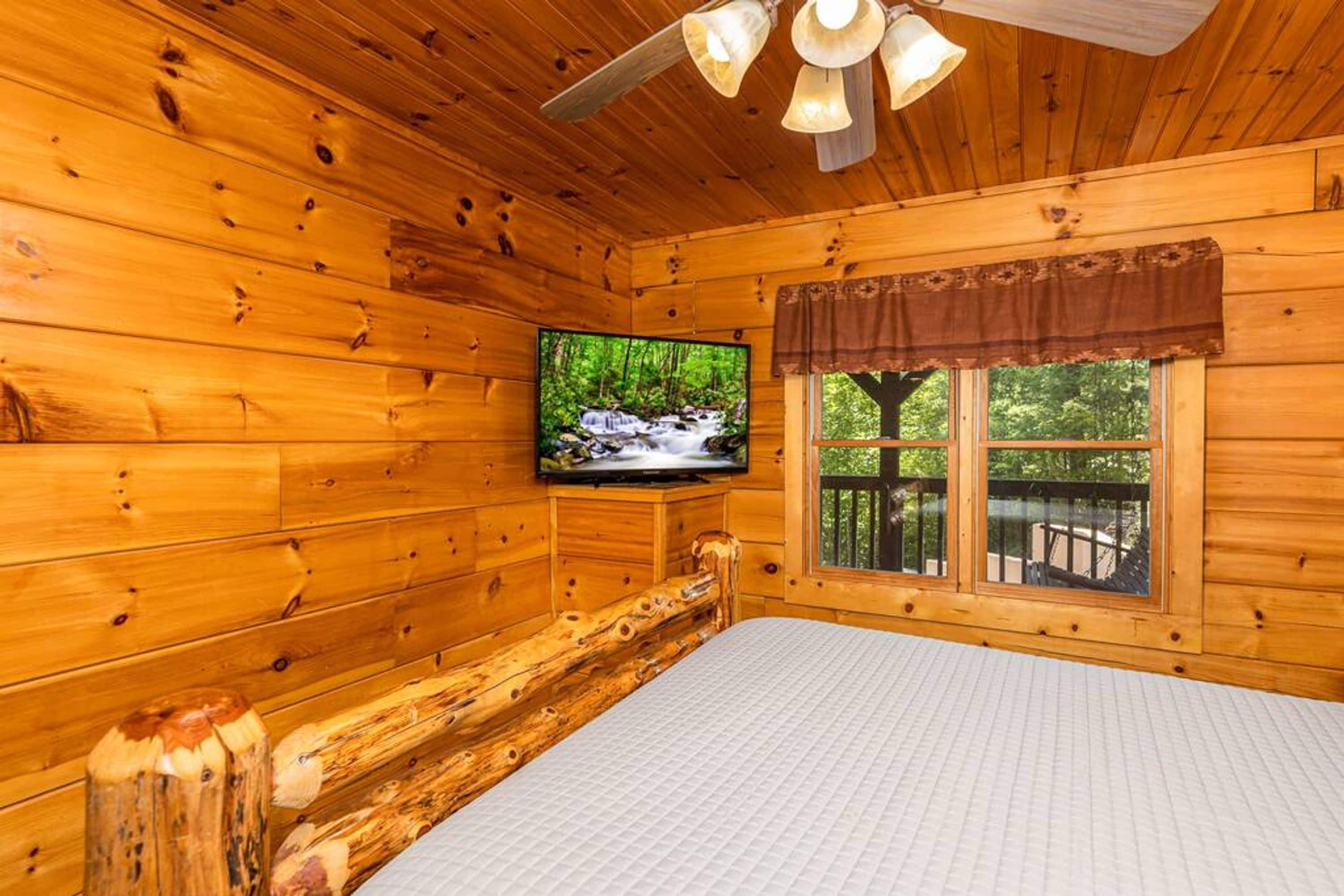 Habitación de cabaña de madera con una cama, un televisor montado en la pared y una ventana con vista a follaje.