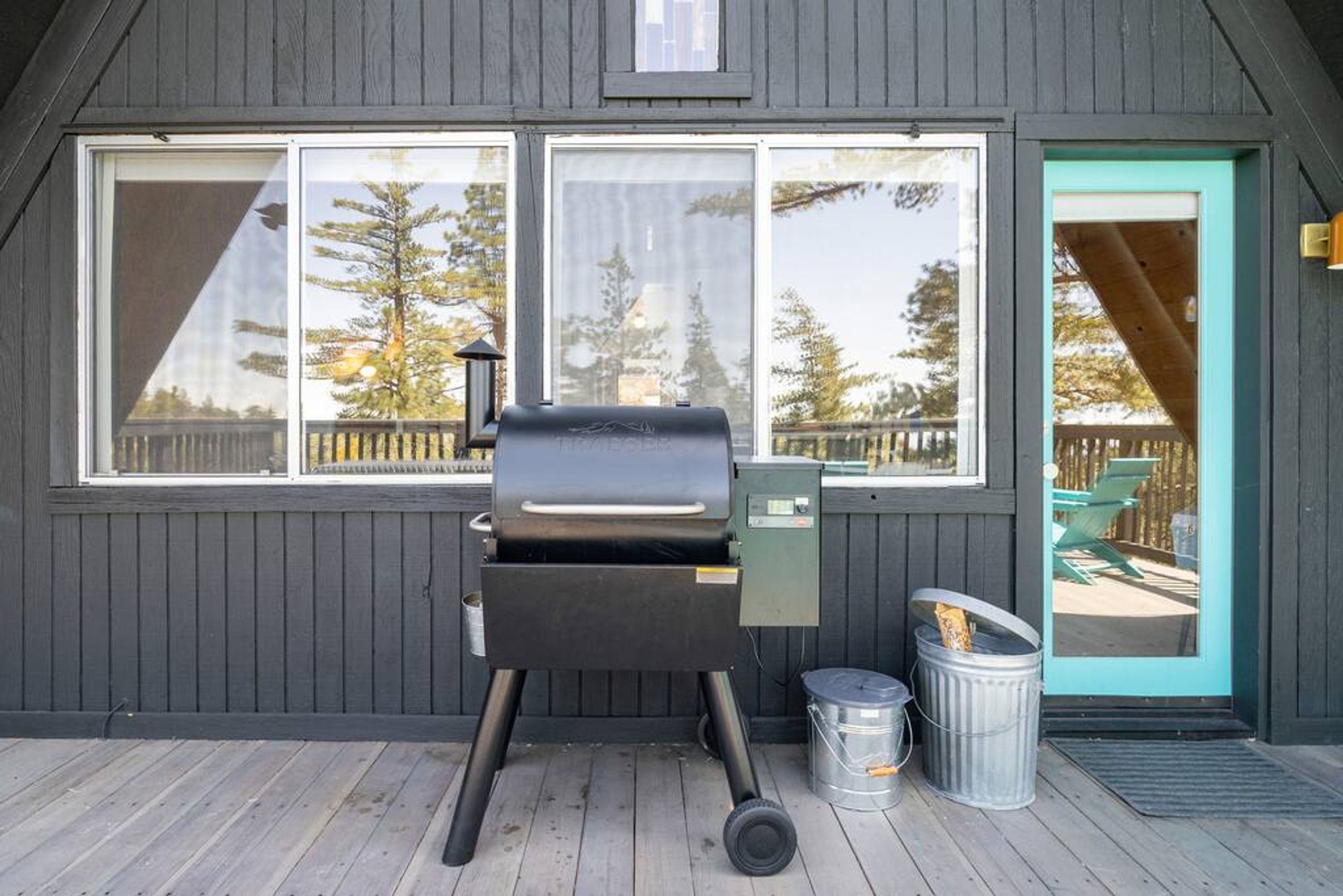 Une terrasse en bois avec un barbecue, une poubelle et une porte donnant sur une autre terrasse et des chaises.