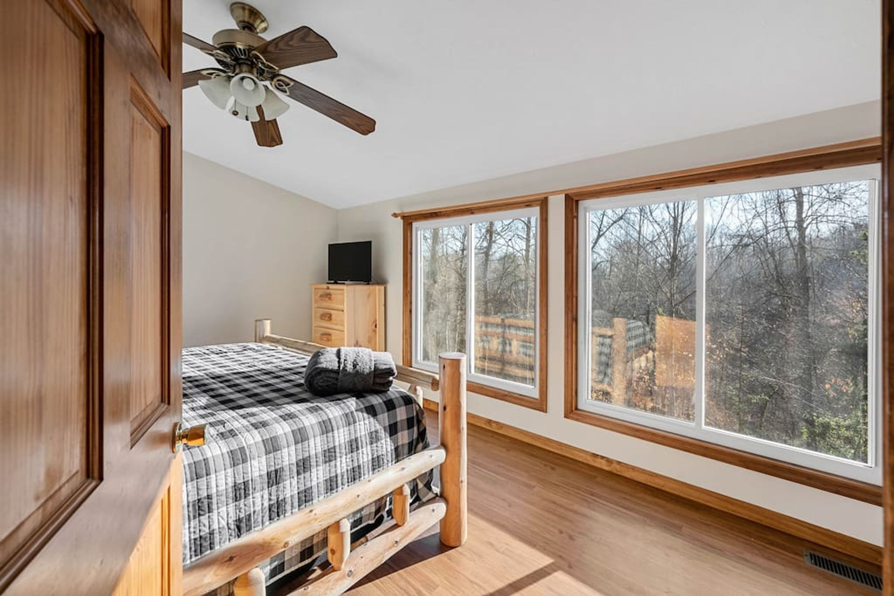 A bedroom with a bed, wooden headboard, a ceiling fan, a dresser, and large windows offering a view of trees.