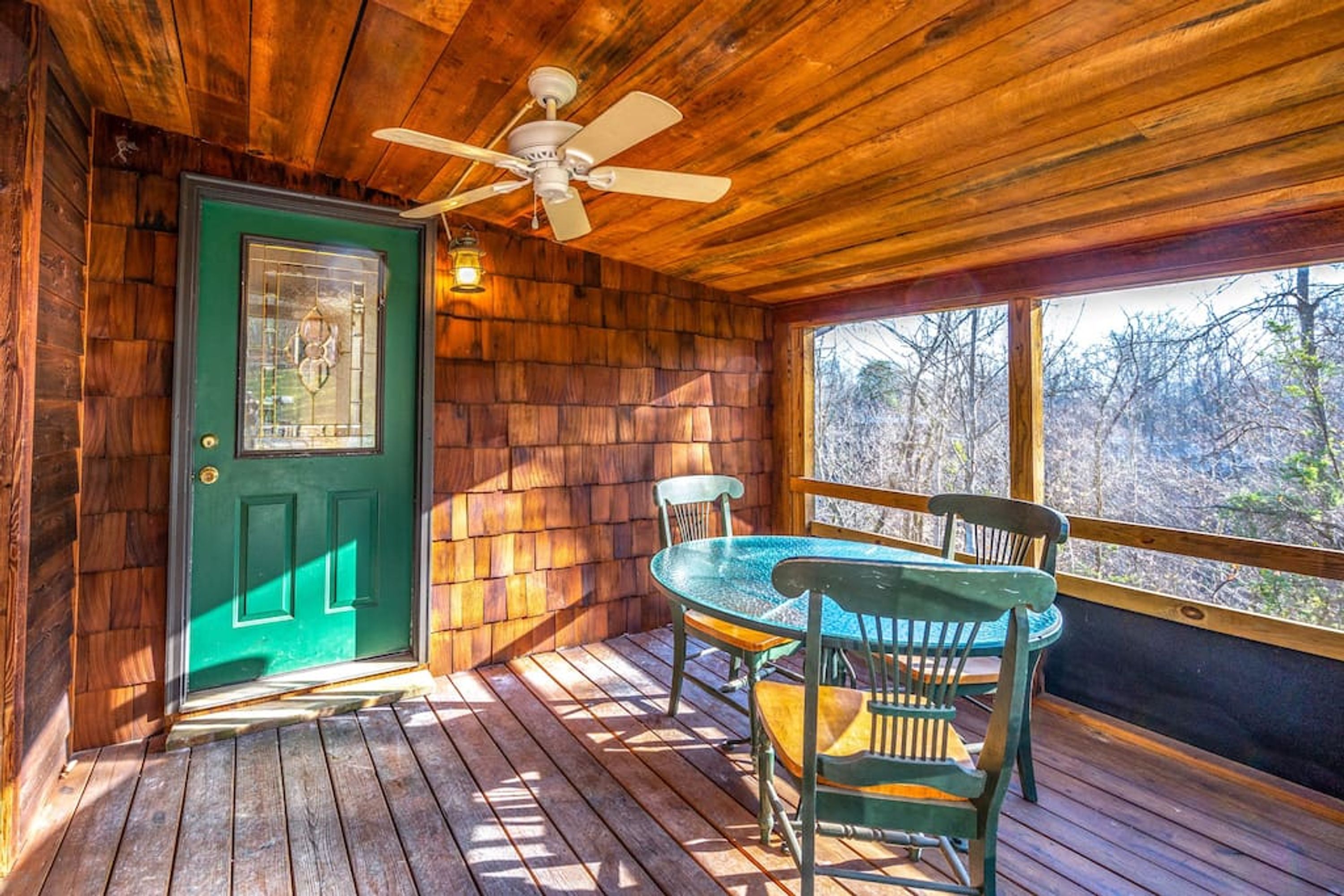 A wooden cabin with a green door, a ceiling fan, a table with chairs, and a view of trees through a large window.