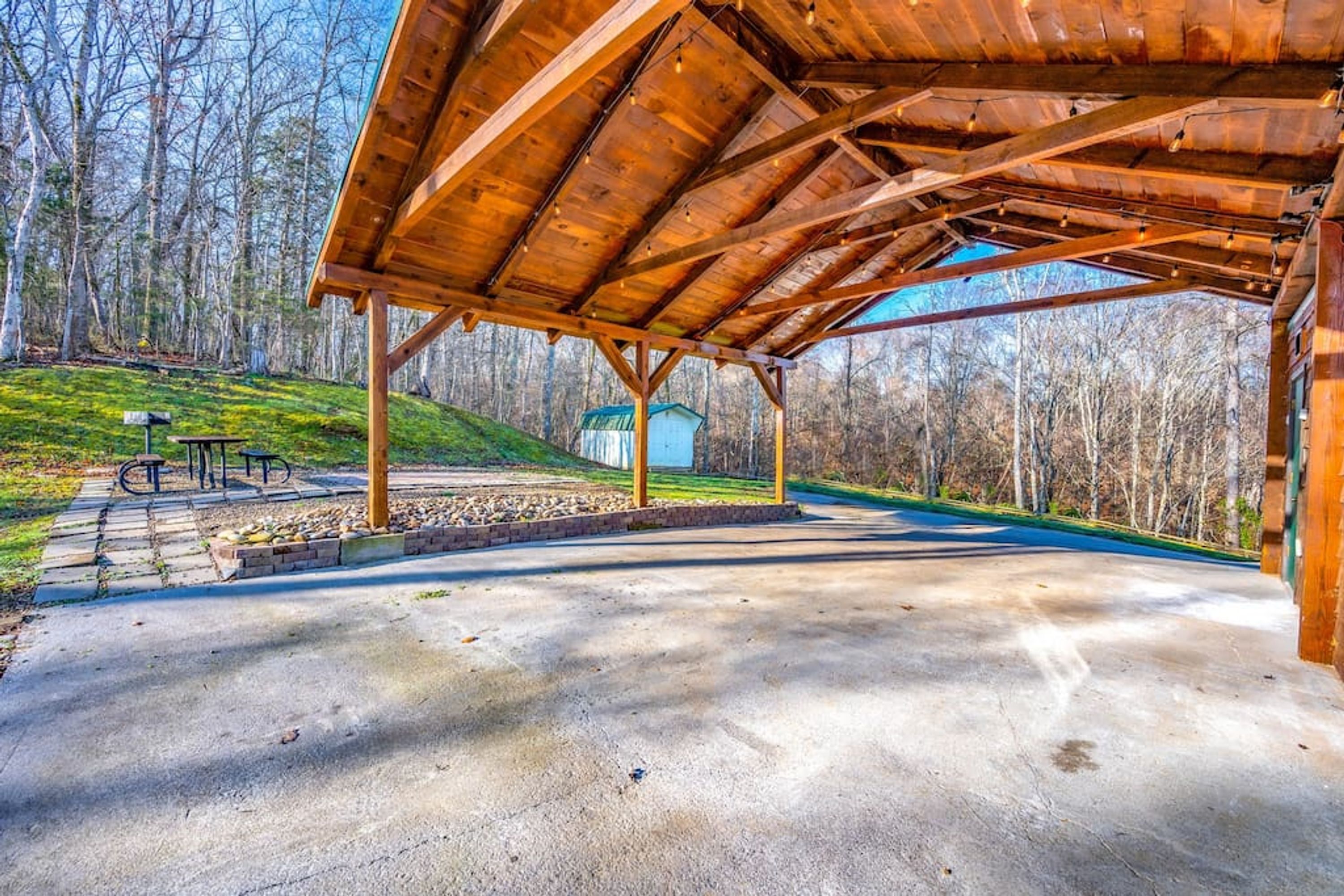 A covered outdoor area with a concrete floor, wooden beams, and a view of a house in the background.