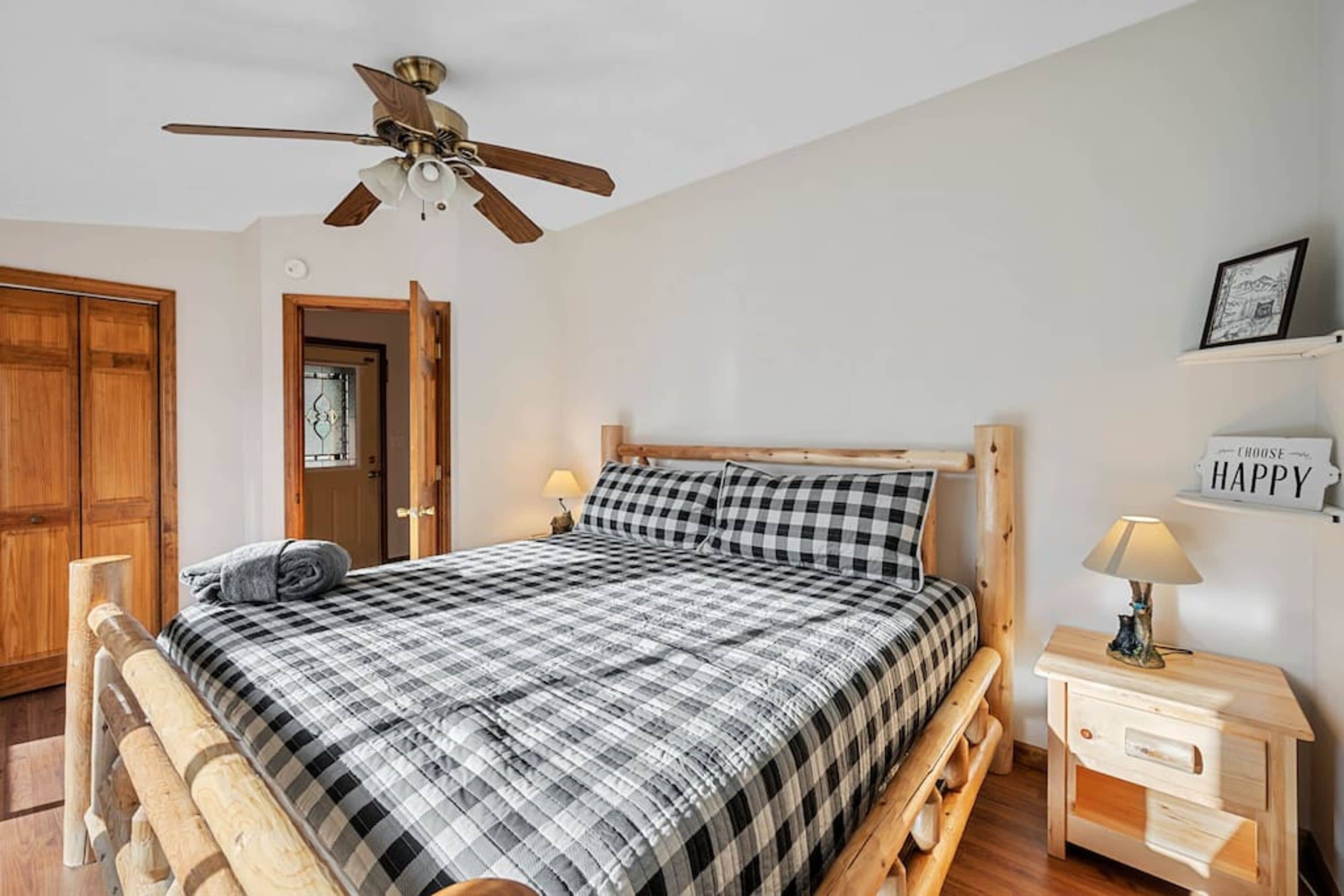 Un dormitorio con cama, cabecera de madera, ventilador de techo, mesita de noche con lámpara y un cartel en la pared.