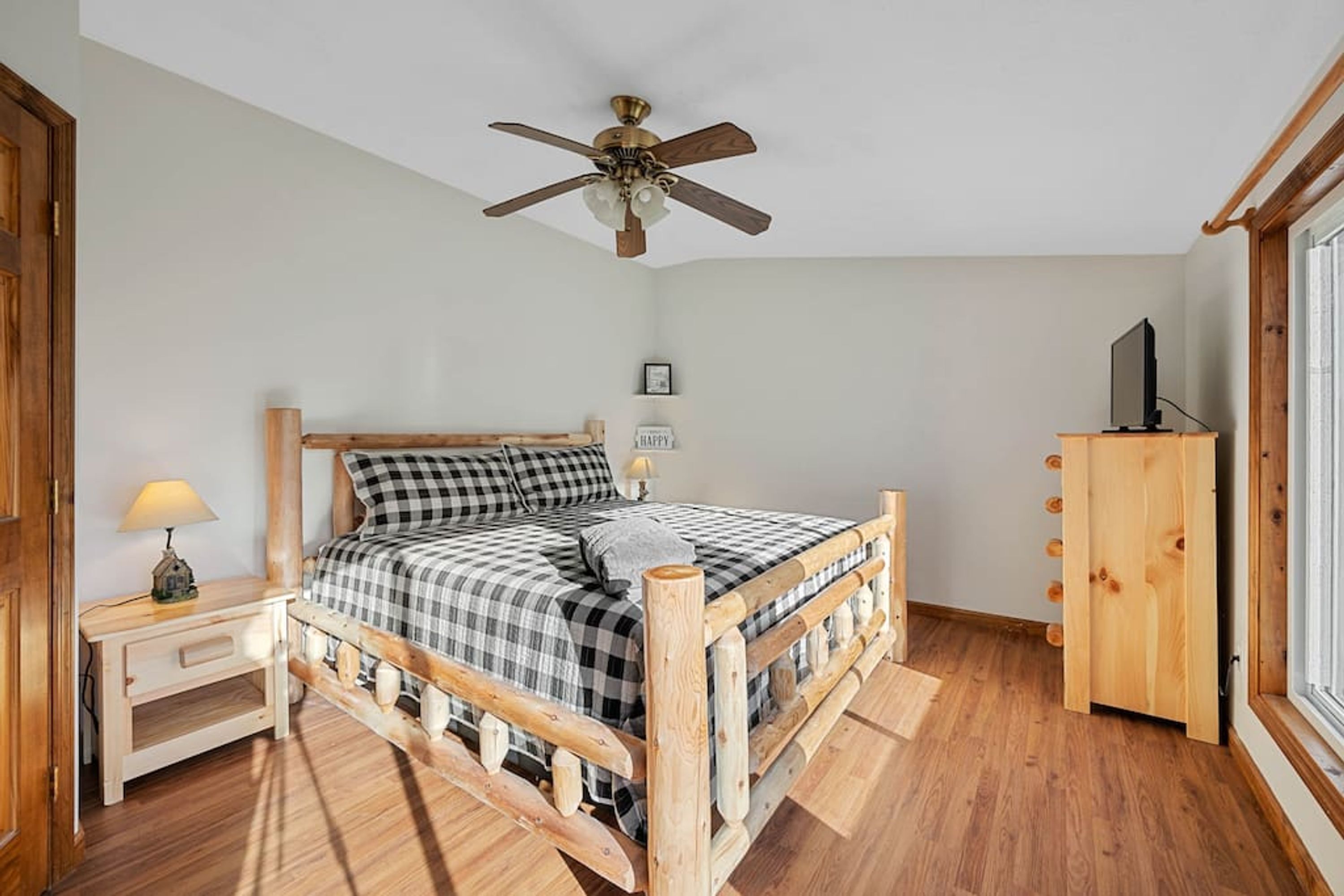 A bedroom with a wooden bed, a nightstand with a lamp, a ceiling fan, a wooden wardrobe, and a window.