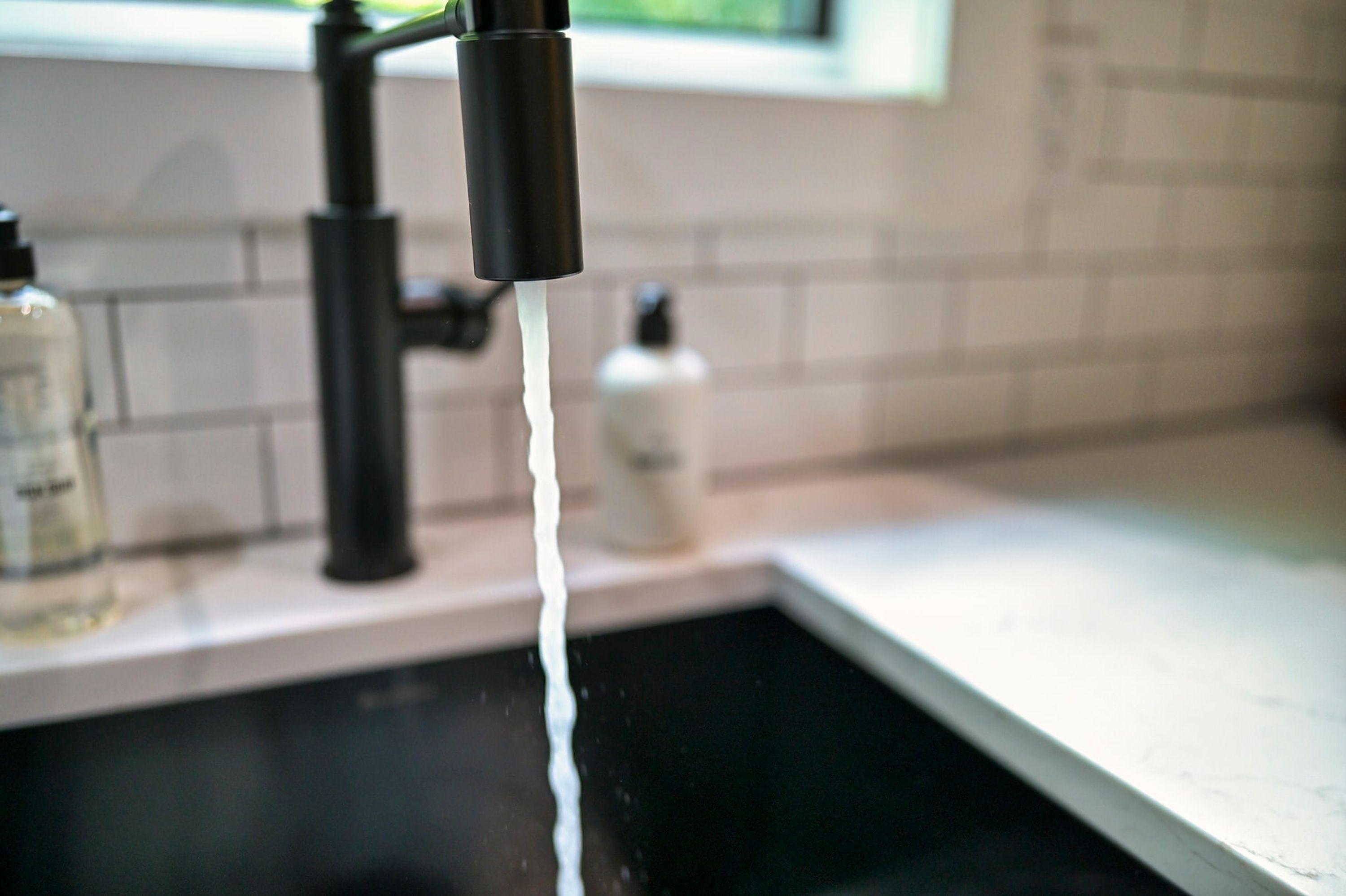 A kitchen sink with running water, a soap dispenser, and a window in the background.