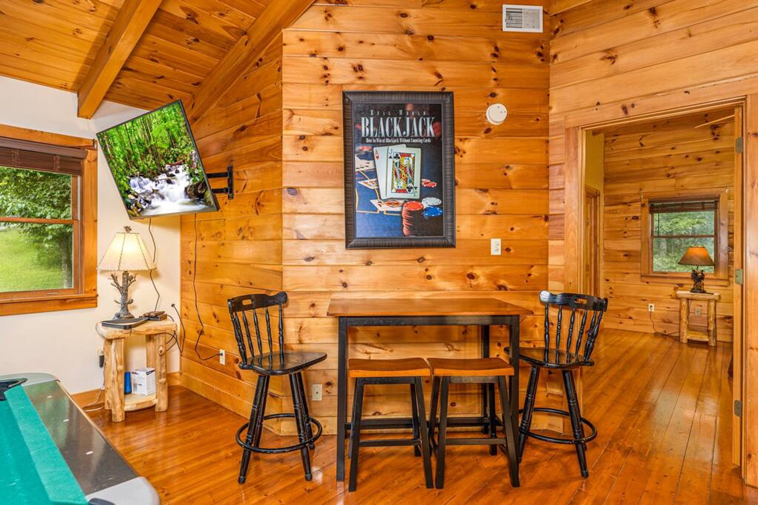 A wooden cabin interior with a dining table, two chairs, a television mounted on the wall, and a framed picture of a blackjack game.