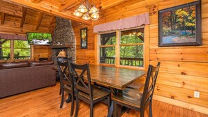 A wooden cabin interior with a dining table, chairs, a sofa, a fireplace, and large windows offering a view of the forest. - Pigeon Forge Cabin Game Room Dog Friendly Hot Tub (Pigeon Forge)