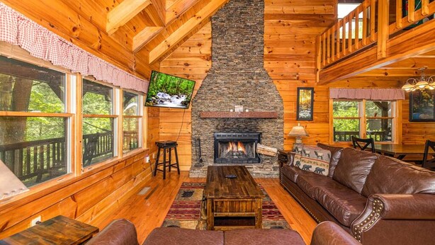 A cozy cabin interior with a stone fireplace, wooden walls, leather seating, and large windows offering a view of the outdoors. - Pigeon Forge Cabin Game Room Dog Friendly Hot Tub (Pigeon Forge)