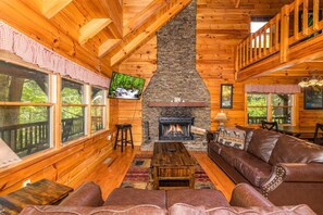 A cozy cabin interior with a stone fireplace, wooden walls, leather seating, and large windows offering a view of the outdoors. - Pigeon Forge Cabin Game Room Dog Friendly Hot Tub (Pigeon Forge)