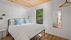 A bedroom with a wooden ceiling, white walls, a bed with white and blue bedding, a bedside table, a hanging light, and a window with a view of greenery. - 2bdrm Cabin 3 Gburg Pet-friendly Firepit Games (Gatlinburg)