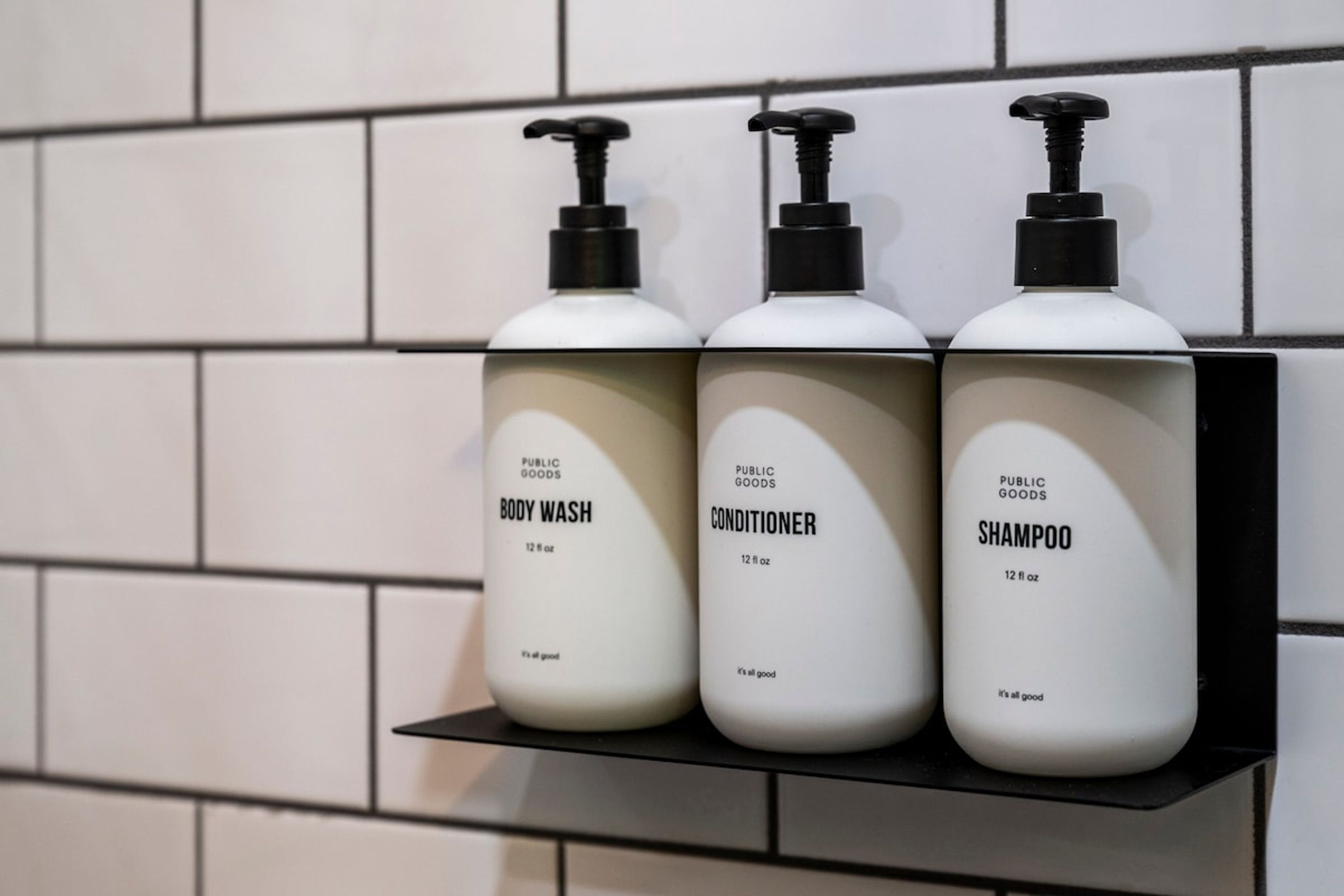 Three bottles of body wash, conditioner, and shampoo on a black shelf against a tiled wall.