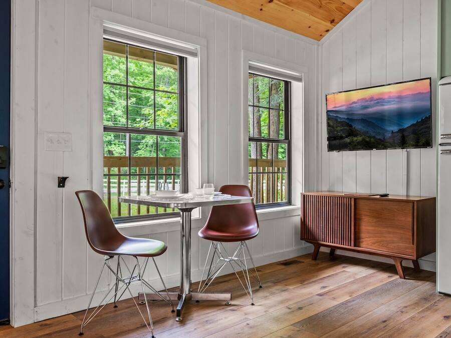 A room with a wooden ceiling, white paneled walls, a dining table with two chairs, a TV on the wall, and a refrigerator.