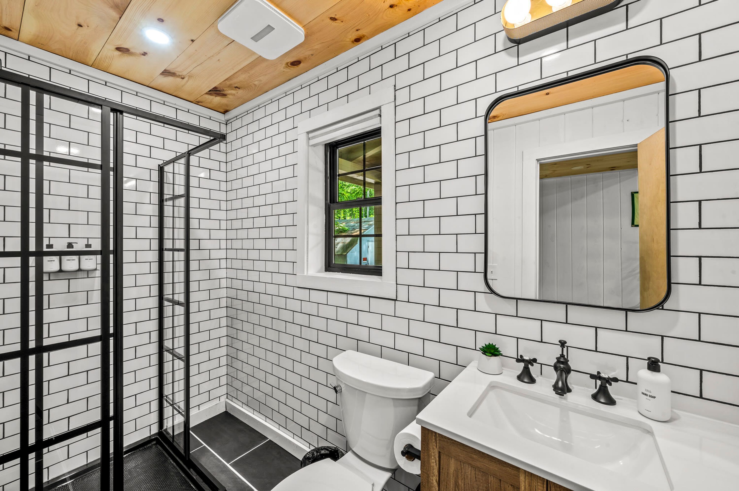 Een moderne badkamer met witte metrotegels, een glazen douchecabine, een wit toilet, een wastafel met zwarte kraanwerk en een raam met uitzicht op groen.