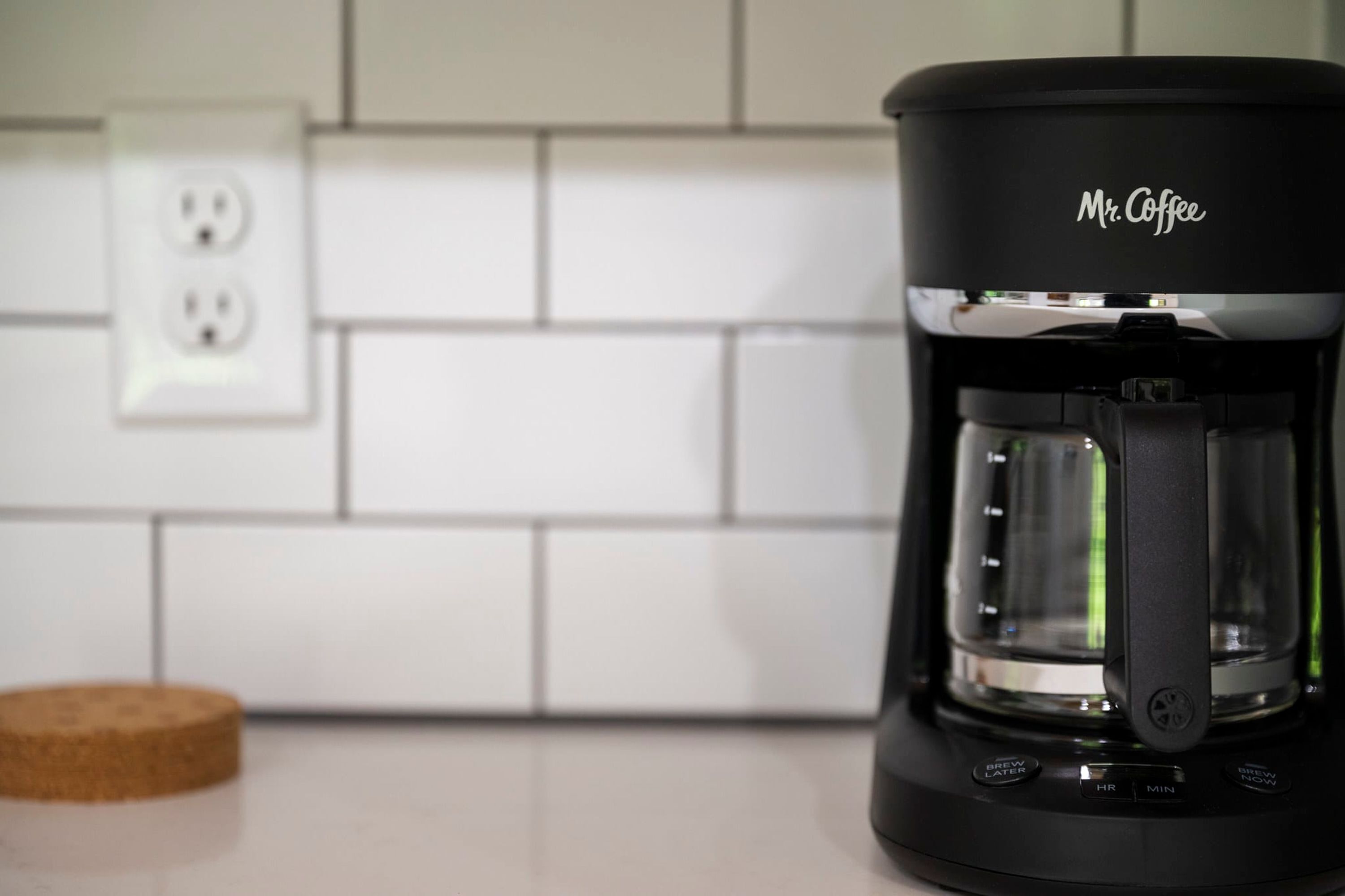 Una cafetera Mr. Coffee negra sobre una encimera de cocina con un salpicadero de azulejos blancos y un posavasos de corcho.