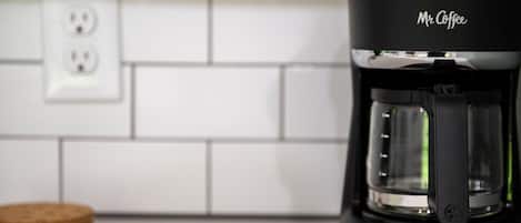 Una cafetera Mr. Coffee negra sobre una encimera de cocina con un salpicadero de azulejos blancos y un posavasos de corcho.