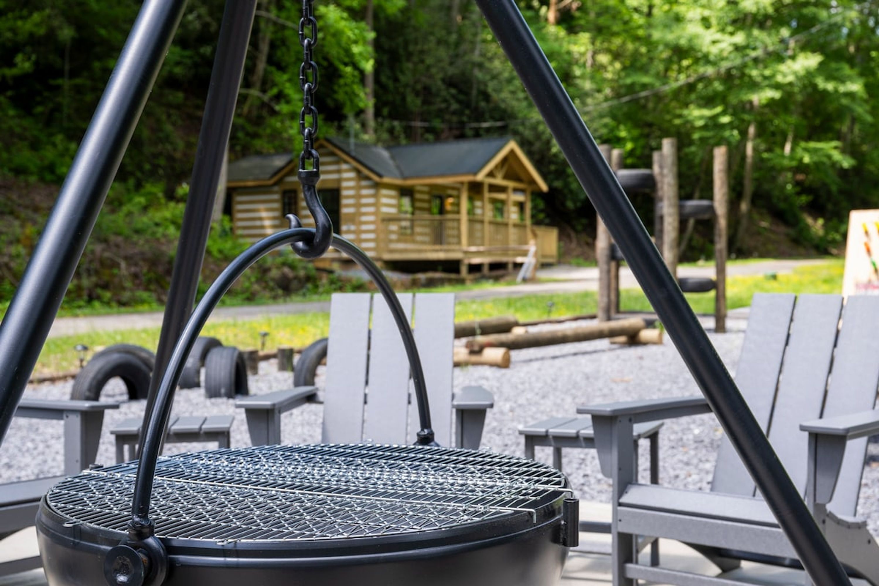 A metal grill suspended over a seating area with wooden chairs and a wooden cabin in the background.