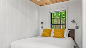 Ein Schlafzimmer mit weißen Wänden, einer Holzbalkendecke, einem Fenster mit Blick auf Bäume, einem Bett mit weißer Bettwäsche und zwei gelben Kissen sowie zwei Nachttischlampen.