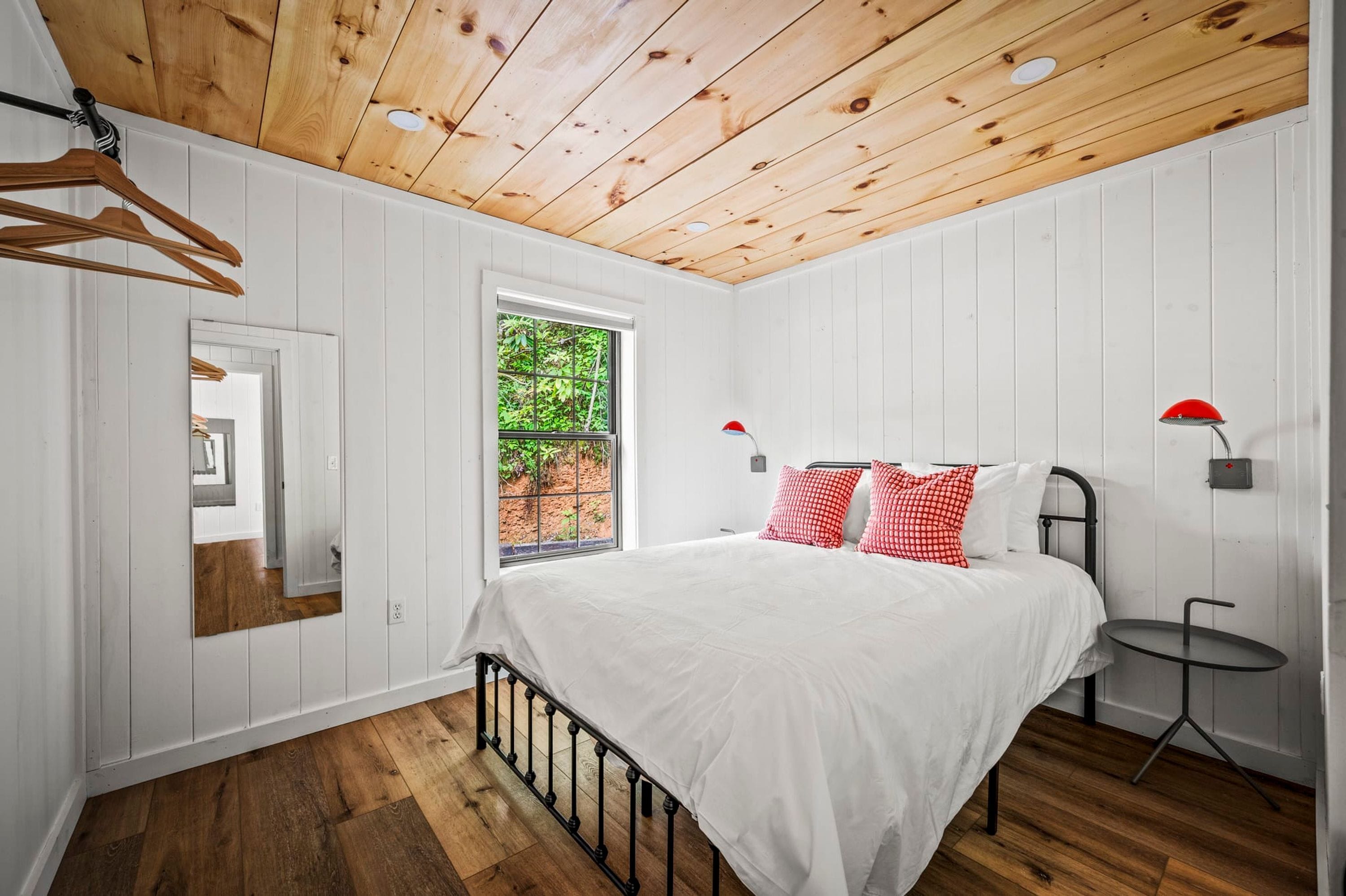 Un dormitorio con techo de madera, paredes blancas, una cama con ropa blanca y almohadas rojas, una mesita y una ventana con vistas a la vegetación.