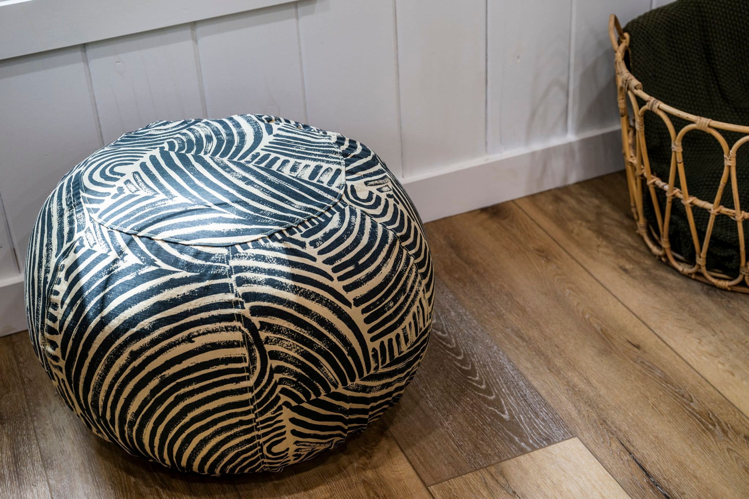A round, patterned ottoman on a wooden floor next to a wicker chair and a white wall.