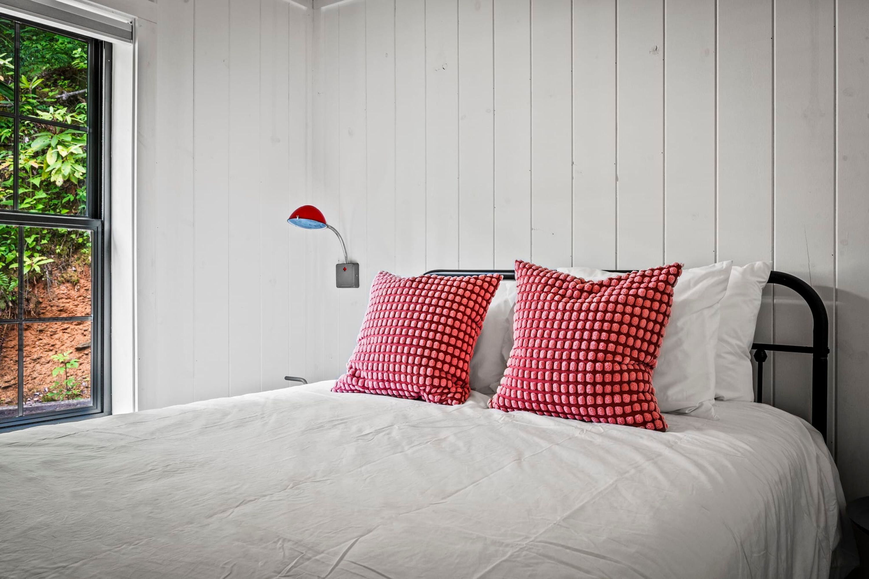 A bedroom with a bed, white bedding, red patterned pillows, a red and blue desk lamp, and a window with a view of greenery.