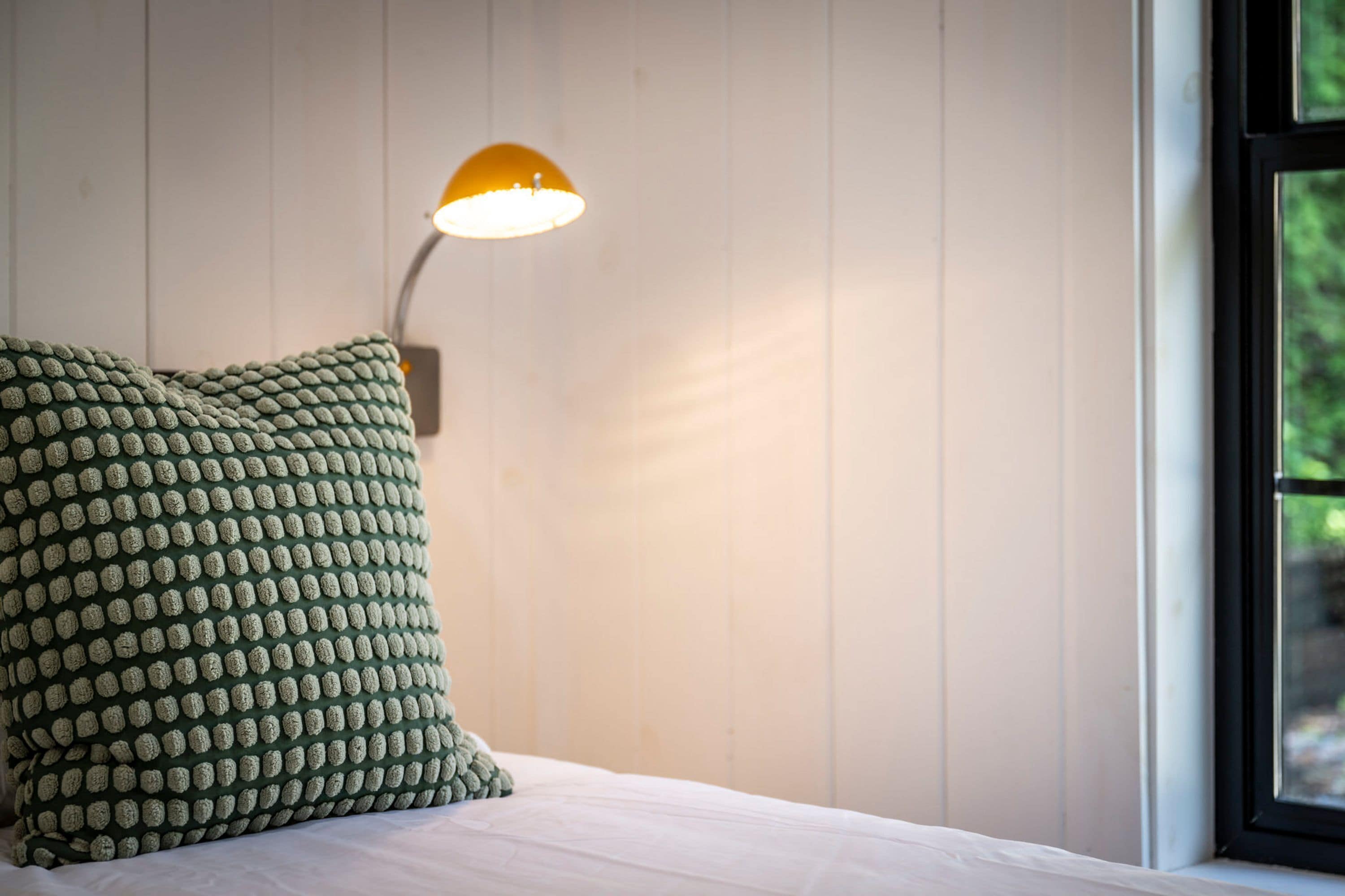 En seng med en grøn pude med et mønster, en lampe på væggen og et vindue med udsigt til grøn vegetation.