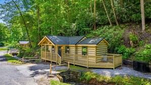 A wooden cabin with a porch and a small deck, surrounded by dense forest.