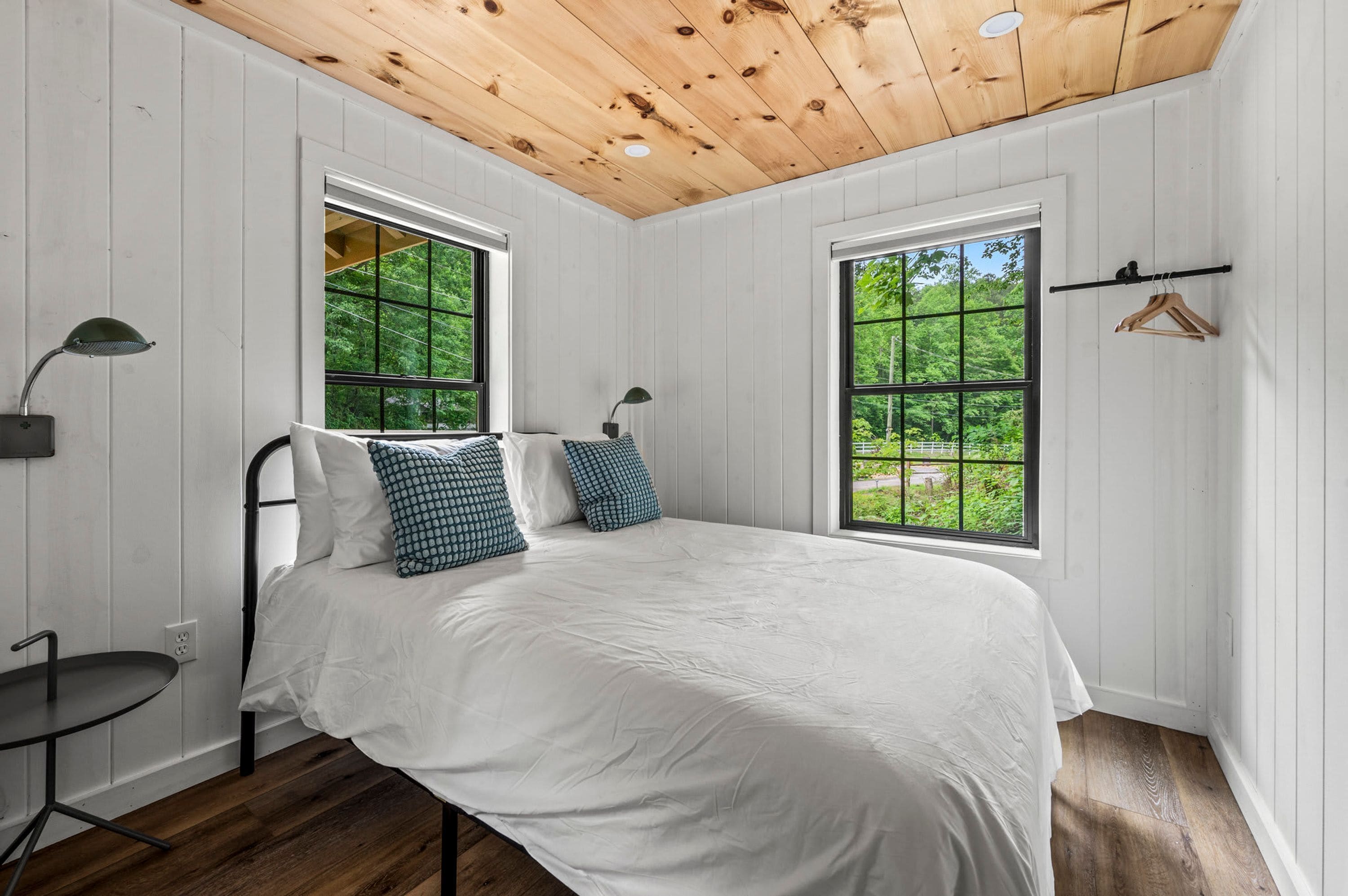 A bedroom with a wooden ceiling, two windows with a view of greenery, a bed with white bedding and patterned pillows, a bedside table, and a hanging light fixture.