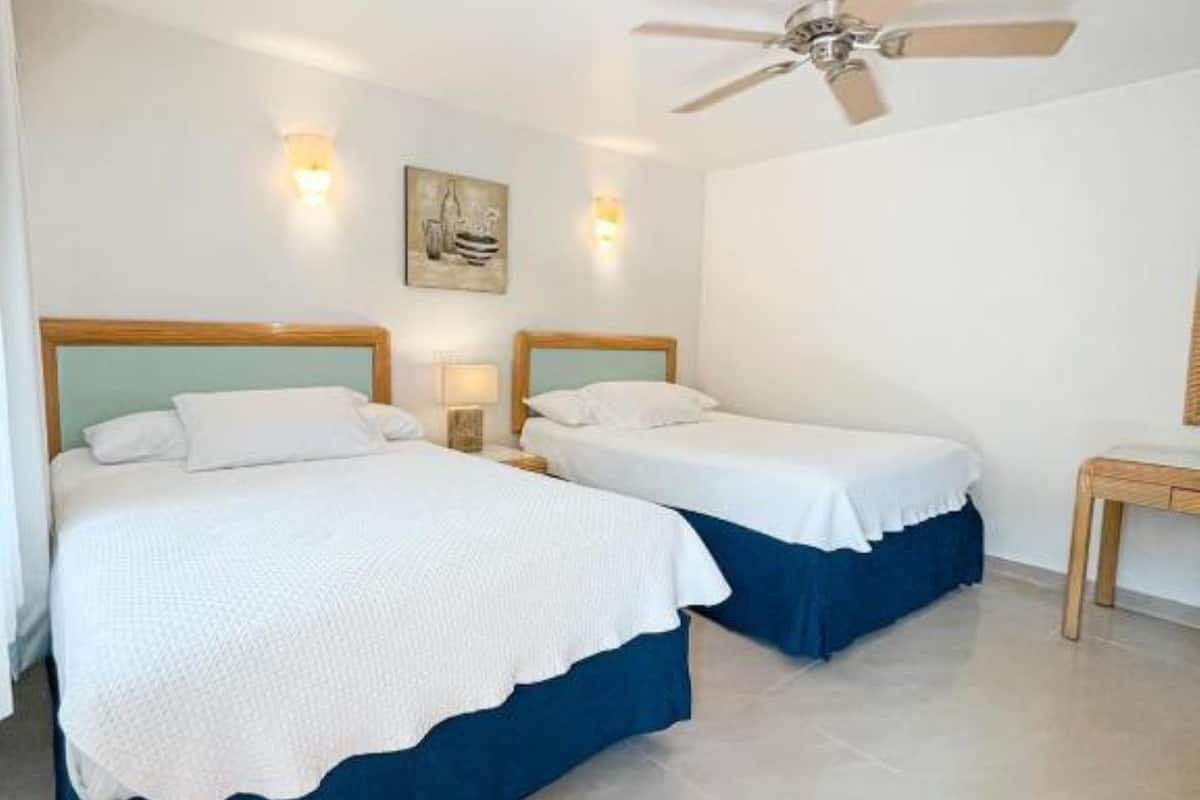Een hotelkamer met twee bedden, een plafondventilator, wandlampen en een klein houten tafeltje.