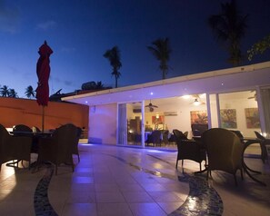 An outdoor dining area with tables and chairs, a red umbrella, and palm trees in the background.