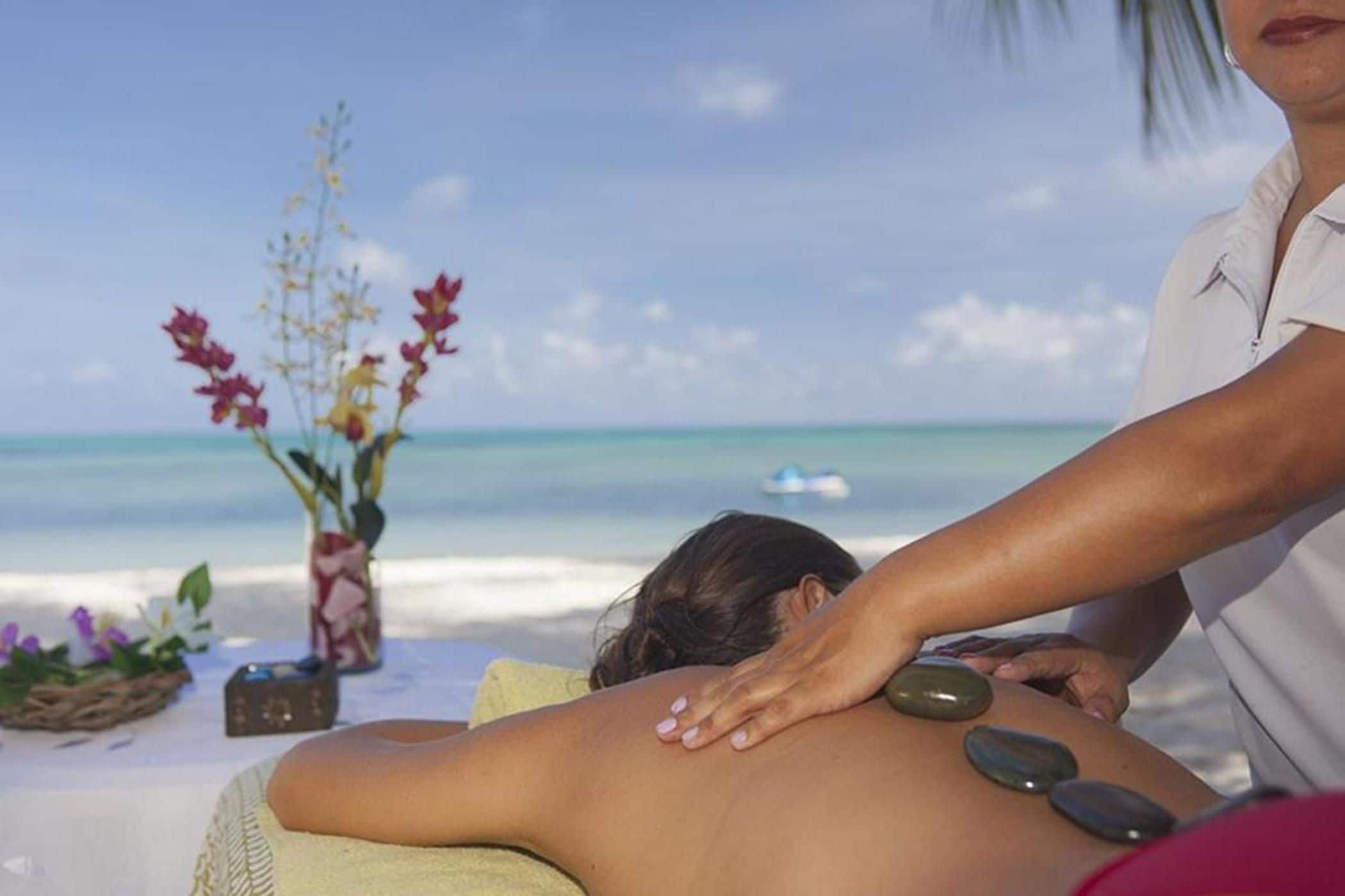 A person receiving a back massage on a beach with a clear view of the ocean and a decorative vase with flowers.