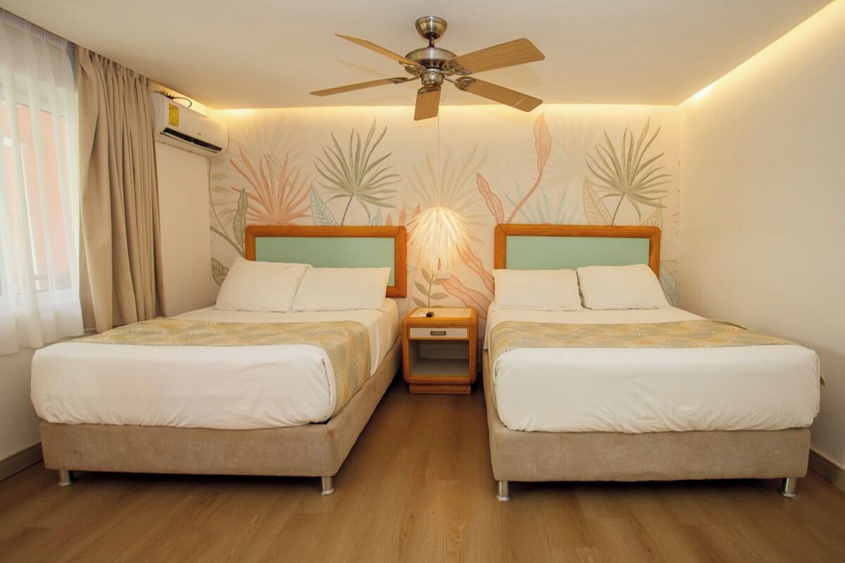 Habitación de hotel con dos camas, un ventilador de techo, una mesita de noche y un ventanal con cortinas.