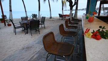 Ein Barbereich direkt am Strand mit Außenbestuhlung, darunter Tische und Stühle, und Blick auf das Meer.