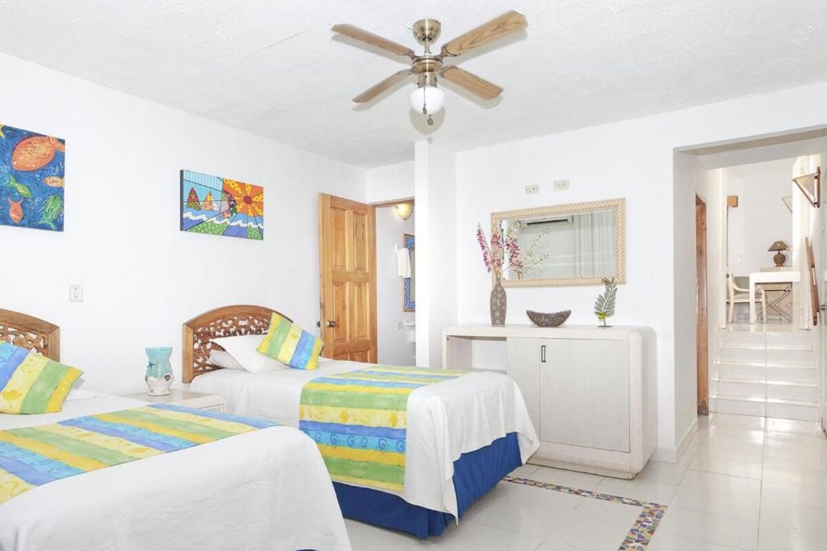 Habitación de hotel con dos camas, un ventilador de techo, un armario blanco y obras de arte en las paredes.