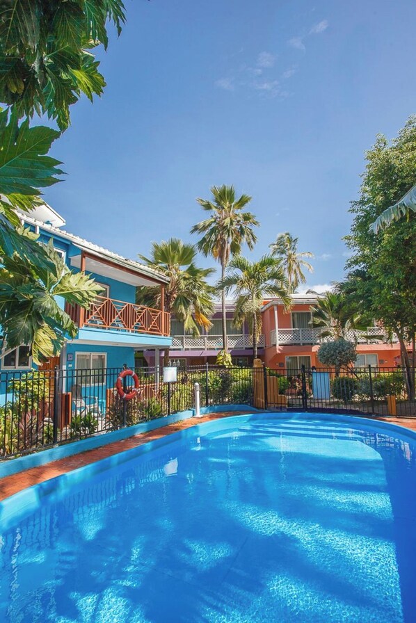 A swimming pool surrounded by buildings with balconies, palm trees, and a clear sky.