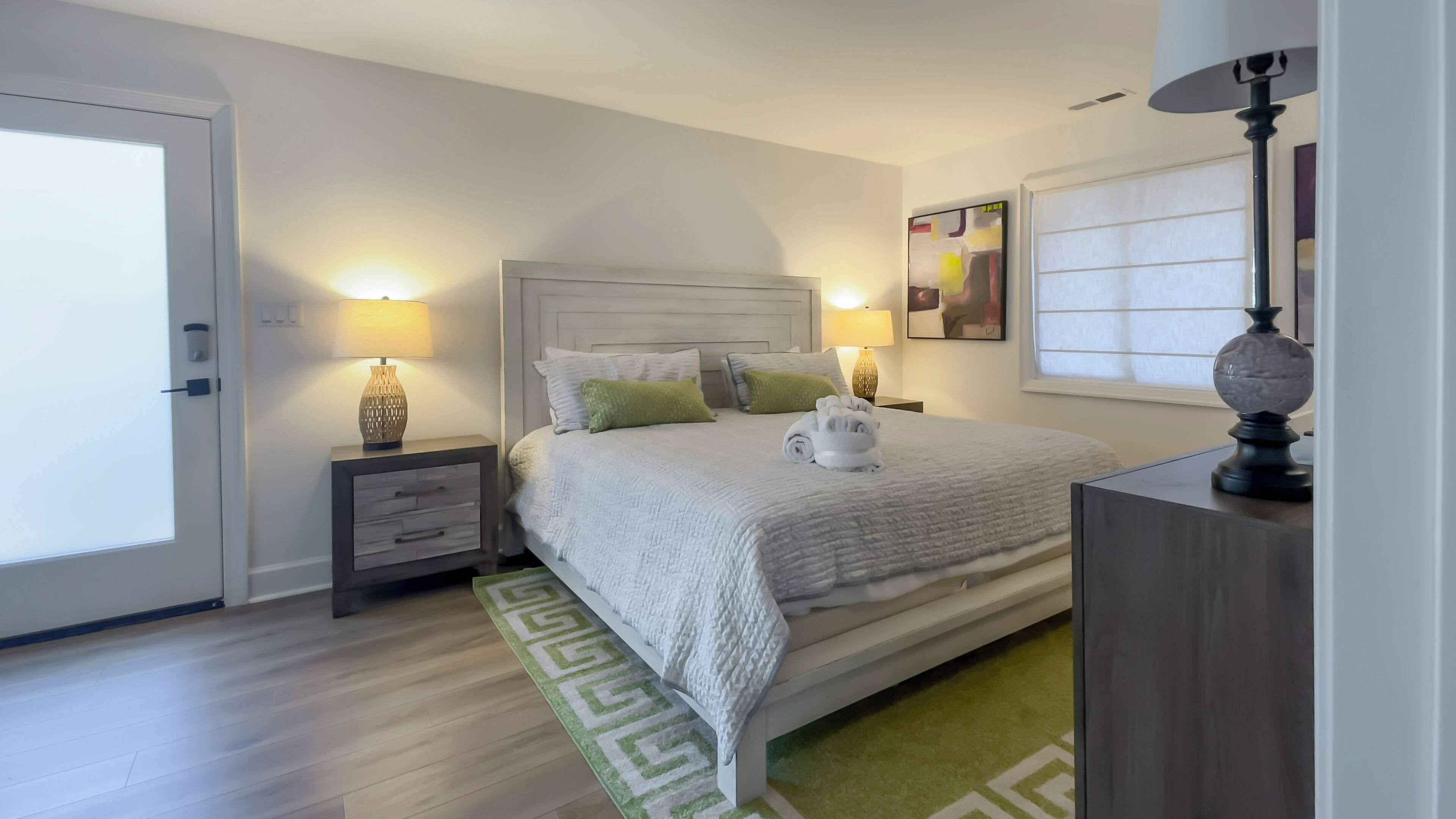 A neatly arranged bedroom with a bed, two bedside tables, a nightstand, a lamp, a painting, and a window with blinds.
