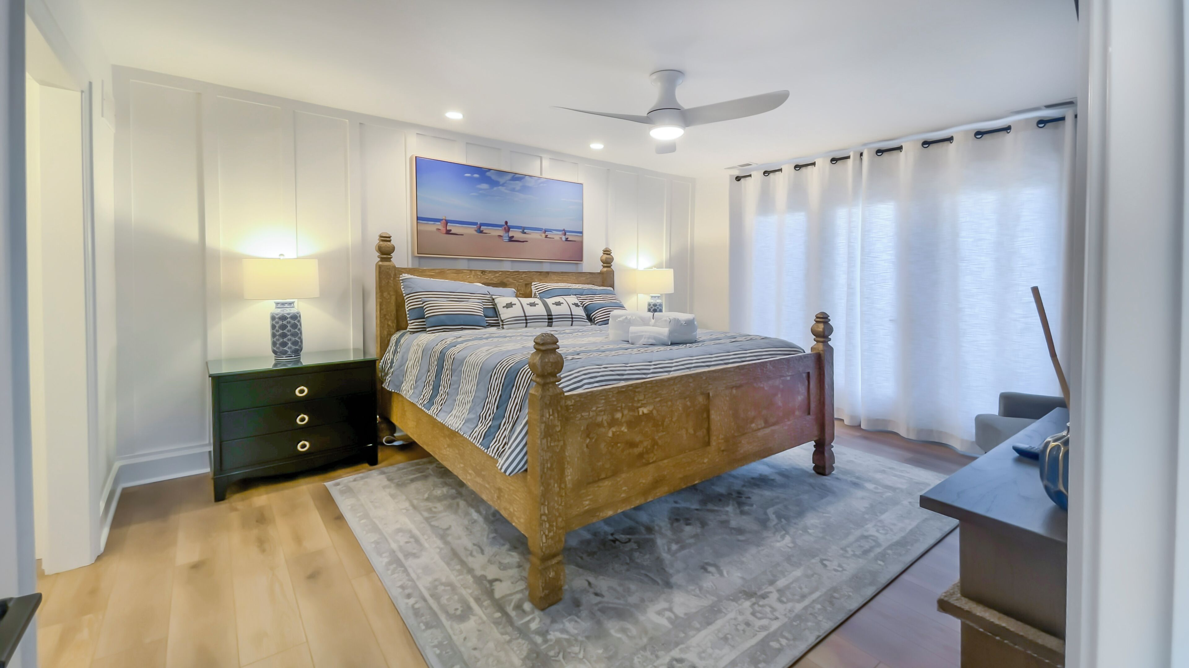 A bedroom with a wooden bed, a nightstand, a desk, a ceiling fan, and a beach-themed painting on the wall.