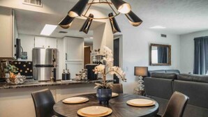 A modern kitchen with a central island, a dining area with a round table and chairs, and a living area with a sofa and a lamp.
