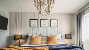 A bedroom with a bed, two bedside tables, a chandelier, a television on the wall, and three framed quotes on the wall.