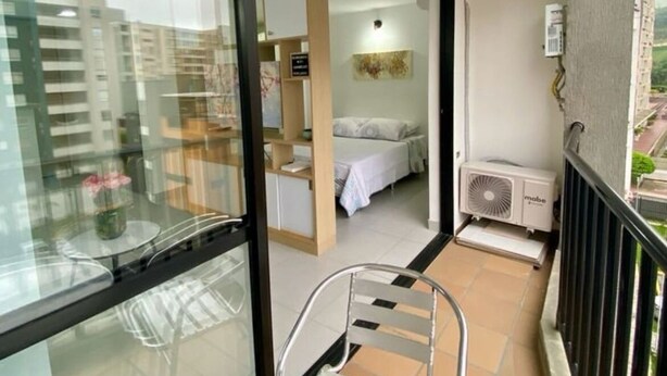 A balcony with a glass door leading to a bedroom. There is a bed with a white blanket, a small table with a chair, and an air conditioning unit on the balcony.