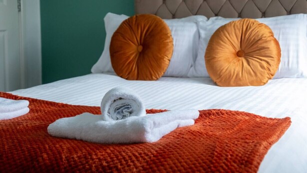 A neatly made bed with white linens, two orange pillows, and a rolled white towel on an orange blanket. - Modern Apartment with Dining, Workspace & Cooking (Motherwell, Glasgow)