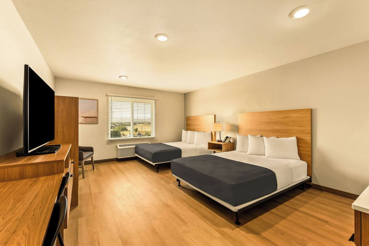 A hotel room with two beds, a desk with a TV, a window with blinds, and a ceiling with recessed lighting.