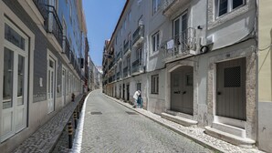 A narrow cobblestone street lined with multi-story buildings, featuring balconies and a person sitting on a chair.