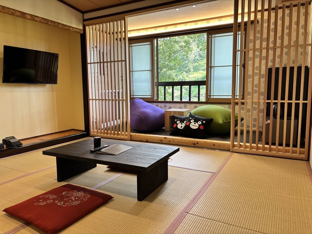 A traditional Japanese room with tatami flooring, a low table, cushions, and a TV mounted on the wall.