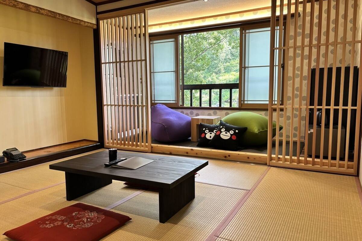 A traditional Japanese room with tatami flooring, a low table, cushions, and a TV mounted on the wall.