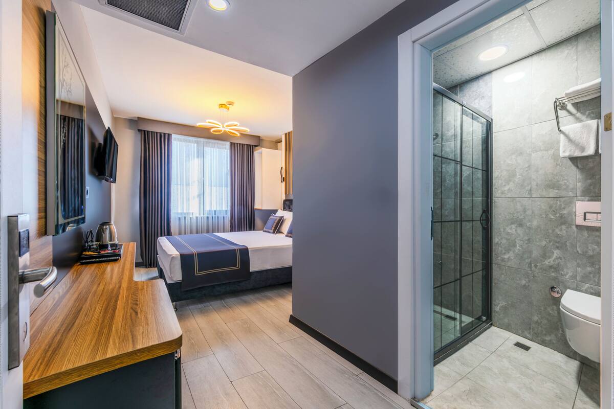 A modern hotel room with a wooden desk, a bed, a glass shower enclosure, and a large mirror.