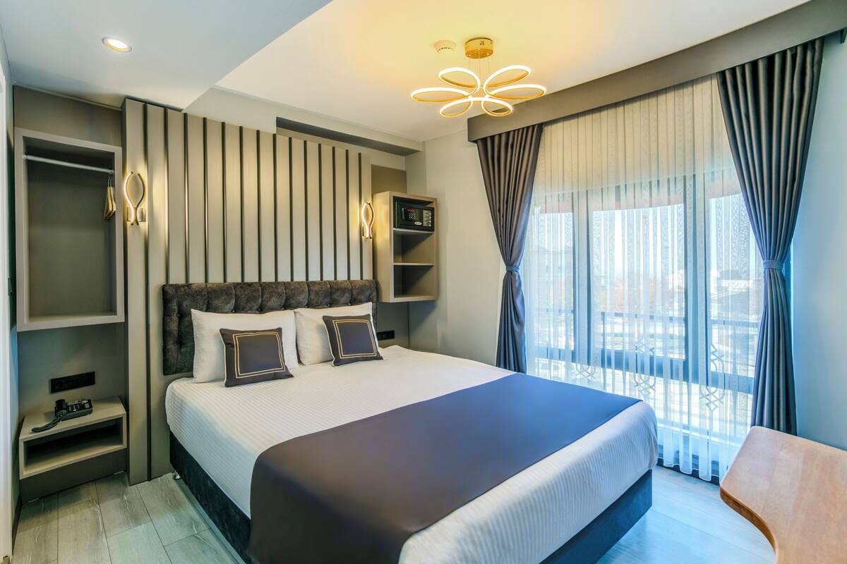 A modern hotel room with a large bed, built-in shelving, a bedside table, and a view of the city through large windows.