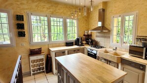Une cuisine dotée d’un grand îlot central, d’une cuisinière, d’un évier et d’un réfrigérateur. On y trouve des fenêtres avec des rideaux, un luminaire suspendu et un meuble contenant des bouteilles de vin.