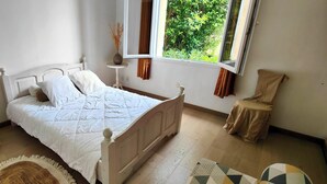 Une chambre à coucher avec un lit, deux fauteuils recouverts de tissu, une table de chevet, une fenêtre donnant sur un paysage verdoyant et un tapis à motifs.