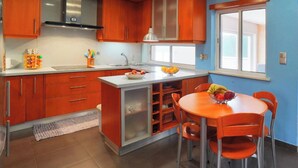 A modern kitchen with wooden cabinets, a stainless steel range hood, a built-in island with a sink, and a dining area with a round table and chairs. - Welcome to SINTRA HOME, a haven between green hills and the blue Atlantic. (São João das Lampas)
